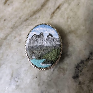 K&ouml;nnte beinhalten: Ovale silberfarbene Brosche mit einer Miniatur-Landschaftsmalerei. Das Gem&auml;lde zeigt schneebedeckte Berge, einen t&uuml;rkisfarbenen Fluss und gr&uuml;nes Laub unter blauem Himmel. Die Brosche hat einen strukturierten, seilartigen Rand.