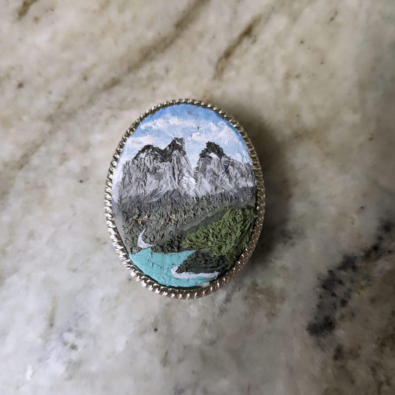 K&ouml;nnte beinhalten: Ovale silberfarbene Brosche mit einer Miniatur-Landschaftsmalerei. Das Gem&auml;lde zeigt schneebedeckte Berge, einen t&uuml;rkisfarbenen Fluss und gr&uuml;nes Laub unter blauem Himmel. Die Brosche hat einen strukturierten, seilartigen Rand.
