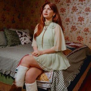 Puede incluir: Una persona con un vestido verde claro con mangas transparentes est&aacute; sentada en una cama con una colcha y almohadas estampadas. Lleva botas blancas y marrones. La habitaci&oacute;n tiene papel pintado floral y una est&eacute;tica vintage.