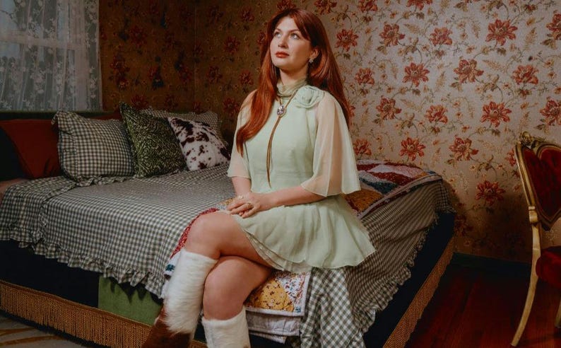 Puede incluir: Una persona con un vestido verde claro con mangas transparentes est&aacute; sentada en una cama con una colcha y almohadas estampadas. Lleva botas blancas y marrones. La habitaci&oacute;n tiene papel pintado floral y una est&eacute;tica vintage.
