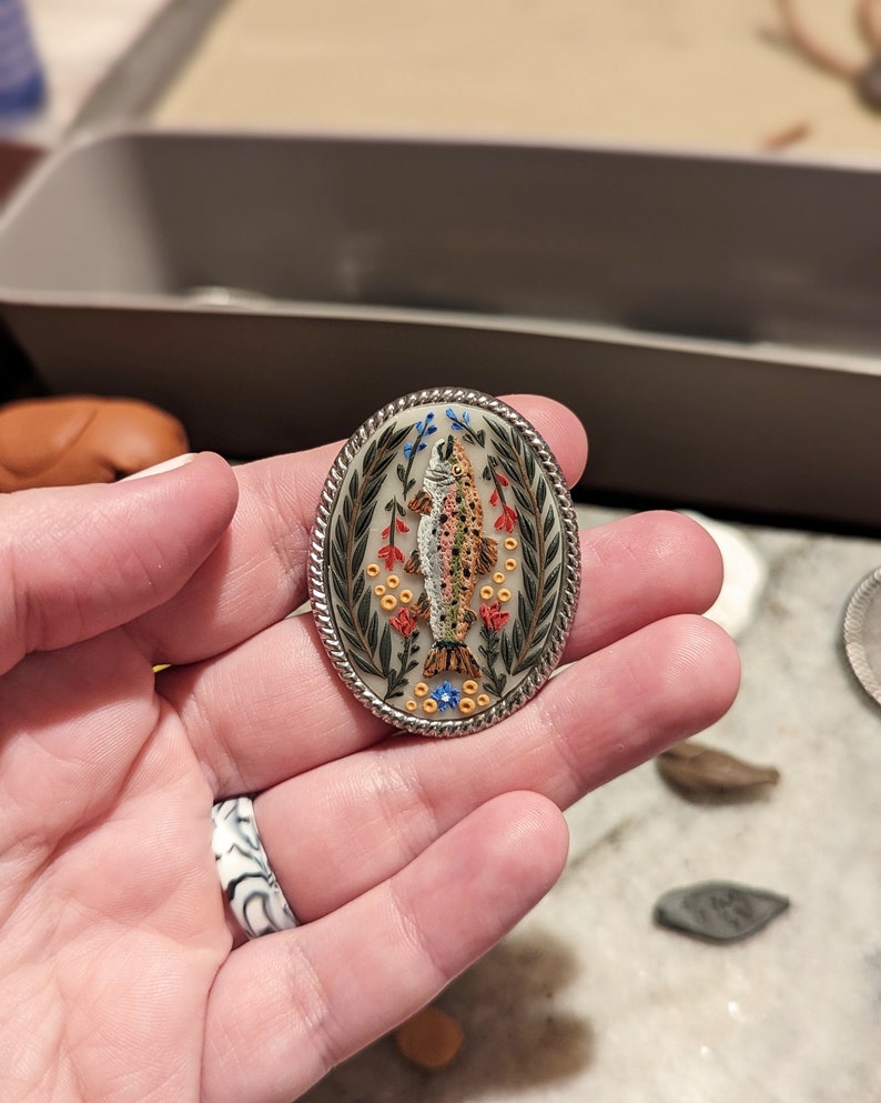 K&ouml;nnte beinhalten: Eine ovale Brosche aus Silber mit einem detaillierten Bild eines Fisches, umgeben von gr&uuml;nen Bl&auml;ttern und roten und gelben Blumen.