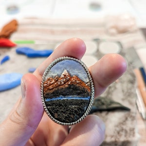 K&ouml;nnte beinhalten: Eine ovale Brosche aus Silber mit einer gemalten Landschaftsszene mit Bergen, einem Fluss und einem bew&ouml;lkten Himmel.