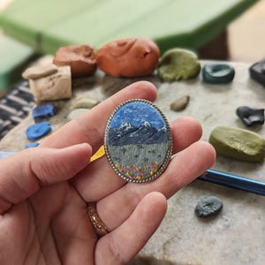 K&ouml;nnte beinhalten: Ovaler Anh&auml;nger mit einer Berglandschaft. Der Anh&auml;nger zeigt einen blauen Himmel mit schneebedeckten Bergen, ein graues Feld und bunte Blumen. Er ist in einen silberfarbenen Metallrahmen mit einem seilartigen Rand gefasst.
