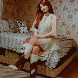 Puede incluir: Una mujer en una habitaci&oacute;n de estilo vintage est&aacute; sentada en una cama con papel tapiz floral. Lleva un vestido verde claro con capa, botas blancas y calentadores de piernas marrones y blancos. La cama tiene un patr&oacute;n de cuadros.
