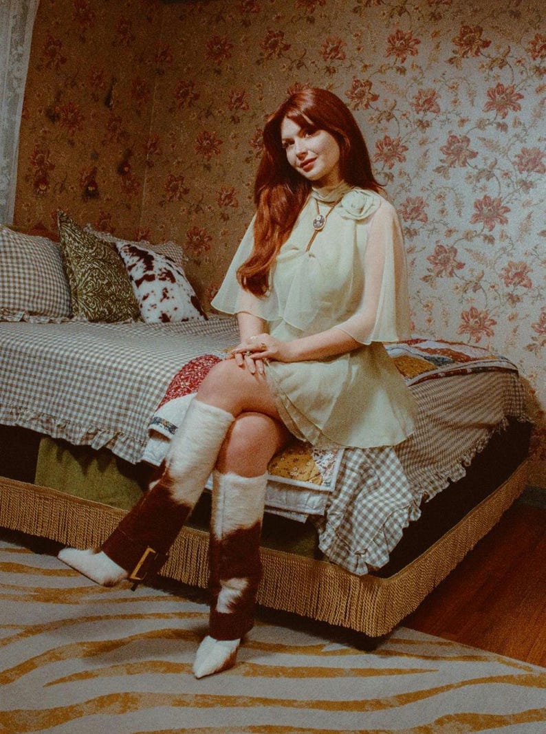 Puede incluir: Una mujer en una habitaci&oacute;n de estilo vintage est&aacute; sentada en una cama con papel tapiz floral. Lleva un vestido verde claro con capa, botas blancas y calentadores de piernas marrones y blancos. La cama tiene un patr&oacute;n de cuadros.