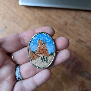 K&ouml;nnte beinhalten: Ovale Brosche mit einer detaillierten Landschaftsszene. Die Brosche zeigt einen blauen Himmel mit wei&szlig;en Wolken, eine braune Felsformation und gr&uuml;ne B&auml;ume. Die Landschaft ist von einem silberfarbenen, seiltexturierten Rand umgeben.