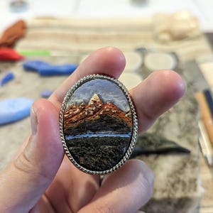 K&ouml;nnte beinhalten: Ovale Brosche mit einer Miniatur-Berglandschaft und einem silberfarbenen, seilartigen Rand. Die Berge sind braun und orange, mit einem wei&szlig;en Gipfel. Ein blauer See und ein dunkler Vordergrund vervollst&auml;ndigen die Szene. Ein Schmuckst&uuml;ck.