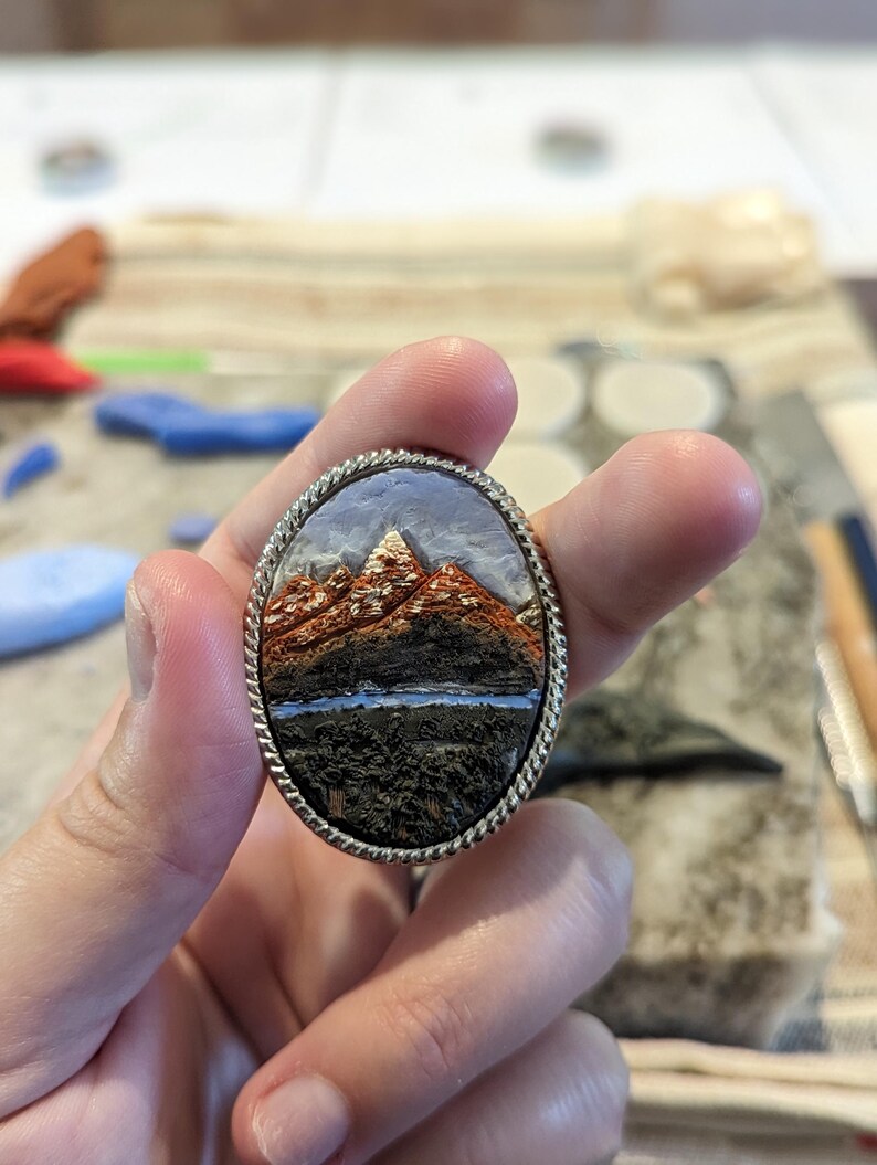 K&ouml;nnte beinhalten: Ovale Brosche mit einer Miniatur-Berglandschaft und einem silberfarbenen, seilartigen Rand. Die Berge sind braun und orange, mit einem wei&szlig;en Gipfel. Ein blauer See und ein dunkler Vordergrund vervollst&auml;ndigen die Szene. Ein Schmuckst&uuml;ck.