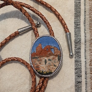 K&ouml;nnte beinhalten: Braune Leder-Bolo-Krawatte mit einem ovalen Anh&auml;nger. Der Anh&auml;nger zeigt eine W&uuml;stenlandschaft mit roten Felsformationen, Kakteen und blauem Himmel. Die geflochtene Lederschnur hat silberfarbene Metallspitzen.