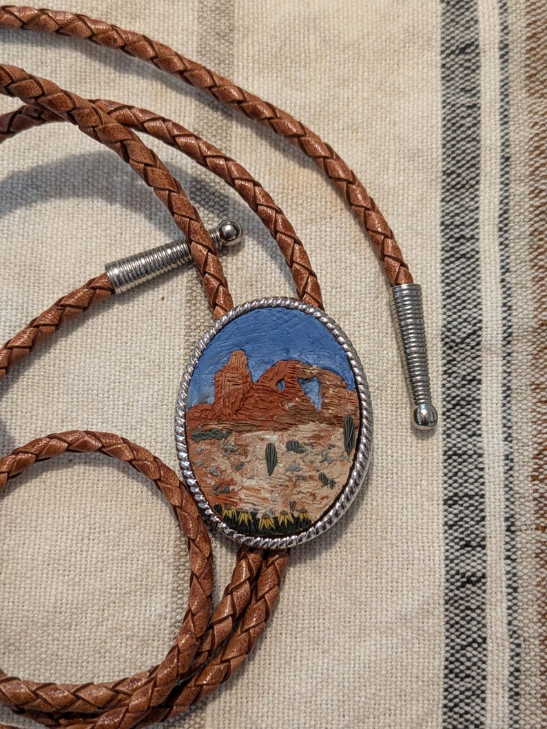 K&ouml;nnte beinhalten: Braune Leder-Bolo-Krawatte mit einem ovalen Anh&auml;nger. Der Anh&auml;nger zeigt eine W&uuml;stenlandschaft mit roten Felsformationen, Kakteen und blauem Himmel. Die geflochtene Lederschnur hat silberfarbene Metallspitzen.