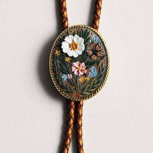 Puede incluir: Corbata bolo ovalada con un dise&ntilde;o floral detallado. La pieza central presenta una flor blanca con centro naranja, una flor rosa y una mariposa. El colgante est&aacute; enmarcado con un borde dorado y cuelga de un cord&oacute;n de cuero marr&oacute;n trenzado.