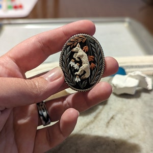 K&ouml;nnte beinhalten: Eine wei&szlig;e Keramik-Fuchsfigur mit orangefarbenen Bl&auml;ttern und wei&szlig;en Bl&uuml;ten, vor schwarzem Hintergrund, ist von einem silberfarbenen Metallrahmen umgeben. Die Figur ist ein detailliertes, handgefertigtes Kunstwerk.