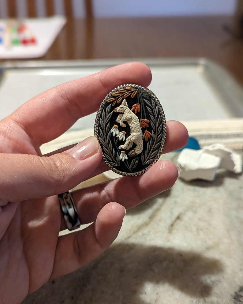 K&ouml;nnte beinhalten: Eine wei&szlig;e Keramik-Fuchsfigur mit orangefarbenen Bl&auml;ttern und wei&szlig;en Bl&uuml;ten, vor schwarzem Hintergrund, ist von einem silberfarbenen Metallrahmen umgeben. Die Figur ist ein detailliertes, handgefertigtes Kunstwerk.