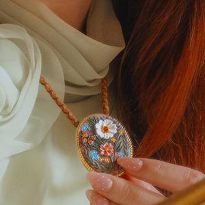 Puede incluir: Un collar con colgante ovalado con un dise&ntilde;o floral. El colgante presenta un marco dorado y un jard&iacute;n en miniatura detallado con flores y mariposas coloridas. El collar tiene un cord&oacute;n de cuero marr&oacute;n.