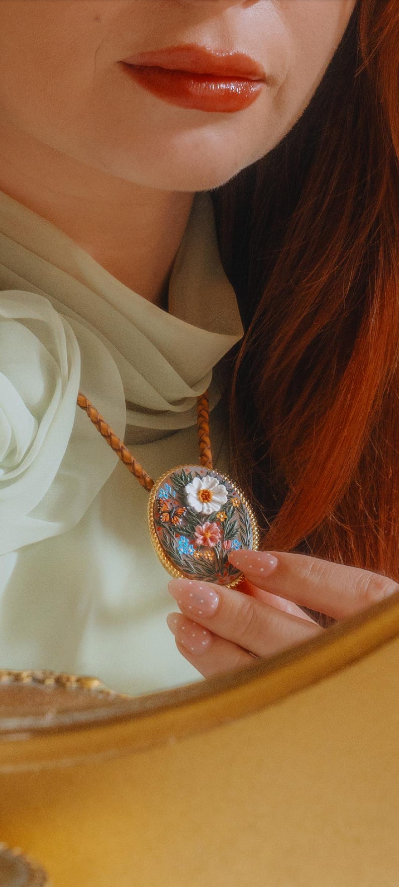 Puede incluir: Un collar con colgante ovalado con un dise&ntilde;o floral. El colgante presenta un marco dorado y un jard&iacute;n en miniatura detallado con flores y mariposas coloridas. El collar tiene un cord&oacute;n de cuero marr&oacute;n.