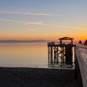 May include: A wooden pier extends out over the water at sunset. The pier has a small shelter at the end and a wooden railing. The water is calm and the sky is a mix of orange, pink, and blue.