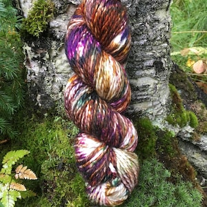 May include: A skein of hand-dyed yarn in shades of brown, purple, white, and green. The yarn is twisted into a spiral shape and is resting on a bed of moss.