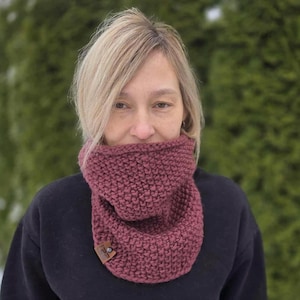 May include: A knitted cowl in a deep burgundy color. The cowl is made of thick yarn, with a textured stitch pattern. A small, brown leather tag with a button is attached to the cowl. The person is wearing a black sweater.
