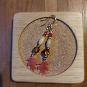 May include: Dangle earrings with an autumnal theme. The earrings have silver hooks, small red beads, light oval beads, and amber glass beads. They are finished with red leaf-shaped charms. The earrings are displayed on a cork coaster.