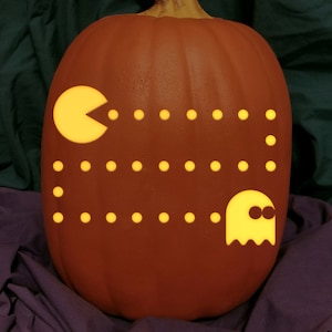May include: A carved orange pumpkin with a Pac-Man design. The design includes Pac-Man, a ghost, and dots, all illuminated with a yellow glow. The pumpkin sits on a purple cloth.