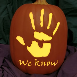 May include: A brown pumpkin with a carved handprint design, illuminated from within with a warm yellow glow. The text "We know" is carved below the handprint. The pumpkin is a Halloween decoration.