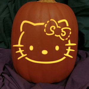 May include: A carved orange pumpkin with a Hello Kitty face design. The design is illuminated from within, creating a glowing effect. The pumpkin has a brown stem and sits on a purple and green fabric background.