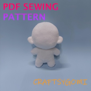 Könnte beinhalten: Eine weiße Plüschpuppenbasis mit einem abgerundeten Kopf, kleinen Ohren und kurzen Gliedmaßen. Der Text PDF SEWING PATTERN ist oben in Pink und Lila und der Text CRAFTSYGOMI unten im Bild in Orange dargestellt.