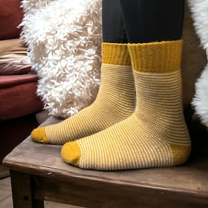 Pode incluir: Meias amarelo mostarda com riscas horizontais brancas. As meias têm punho e biqueira canelados em amarelo mostarda liso. As meias são usadas com leggings pretas. As meias estão numa superfície de madeira.