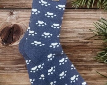 Puede incluir: Calcetines azules con estampado de huellas blancas. Los calcetines, etiquetados como "Jasper's", se describen como "Super Fluffy, Super Soft, Cozy Socks". Se muestran sobre una superficie de madera.