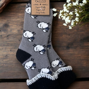 Pode incluir: Par de meias cinzas e aconchegantes com estampa de ovelhas brancas e acabamento preto. As meias têm uma textura felpuda e são etiquetadas como "Super Fluffy Super Soft Cozy Socks" por Jess & Lou.