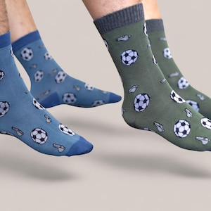 May include: Two pairs of crew socks, one in blue and the other in olive green. Both pairs feature a repeating pattern of soccer balls and whistles. The socks have contrasting colored toes and cuffs.