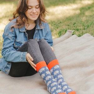 May include: A woman wearing a blue denim jacket and black jeans sits on a beige blanket. She is wearing a pair of blue and orange socks with a white elephant pattern.