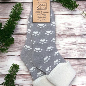Peut inclure: Paire de chaussettes douillettes grises avec des motifs d'empreintes de pattes blanches. Les chaussettes ont un revers blanc moelleux. Les chaussettes sont emballées avec une étiquette brune qui indique "Jess & Lou" et "Super Fluffy Super Soft Cozy Socks".