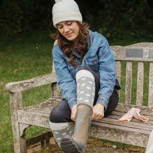 May include: A woman wearing a white knit beanie with a pom-pom, a denim jacket, and gray polka-dot socks. She is sitting on a wooden bench, putting on a pair of rain boots. A pair of pink gloves are on the bench.