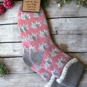 May include: A pair of pink and gray cozy socks with a pattern of gray elephants. The socks have a white fluffy trim around the top. The socks are on a wooden surface with a tag that says "Jess & Lou Super Fluffy Super Soft Cozy Socks".