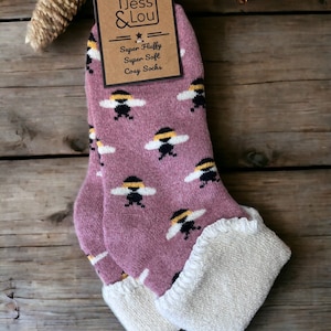 May include: A pair of mauve socks with a repeating bee pattern, featuring a white fuzzy cuff and a cardboard tag labeled Jess & Lou Super Fluffy Super Soft Cosy Socks. The socks have a soft texture and are designed for UK size 4-7.