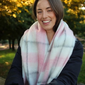 May include: A woman wearing a pink, white, and grey checked scarf. The scarf is wrapped around the neck and shoulders. The woman is smiling and wearing a dark blue jacket. The background is blurred with autumn leaves.