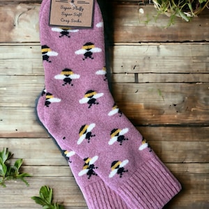 May include: A pair of pink fuzzy socks with a bee pattern. The socks are folded and have a tag that says "Jess & Lou Super Fluffy Super Soft Cozy Socks".