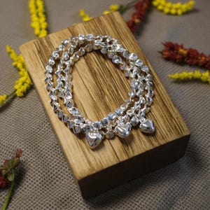 May include: Three silver bracelets with heart charms are displayed on a wooden block. The bracelets have a textured design and are arranged in a circular pattern. The wooden block has a natural wood grain and is surrounded by yellow and red flowers.
