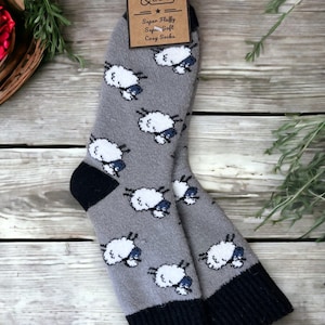 以下が含まれることがあります： 黒いカフと白い羊のデザインが施された灰色の靴下2足。靴下には「Super Fluffy Super Soft Cozy Socks」と「Jess & Lou」と書かれたタグが付いています。