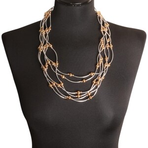 May include: A multi-strand necklace featuring silver-coloured tubes and gold-coloured beads. The necklace is displayed on a black mannequin, highlighting its layered design and metallic accents. The jewellery is designed to be worn around the neck.
