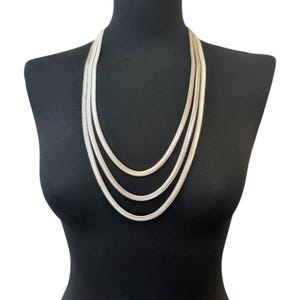 May include: A silver necklace with three strands of different lengths, presented on a black mannequin. The necklace features a flat, snake-chain design, reflecting light. The jewellery is a fashion accessory.