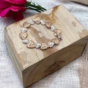May include: A bracelet composed of heart-shaped beads in alternating silver and rose gold tones. The bracelet is displayed on a wooden block, with a pink flower in the background. The hearts have a textured surface.