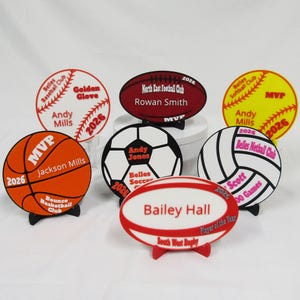May include: A selection of sports awards in various shapes, including round and oval. The awards are for baseball, football, soccer, softball, volleyball, and rugby. Each features team and player details, plus the year 2026.