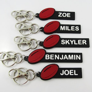 May include: Five black rectangular keychains with silver metal lobster clasps and rings. Each features a raised red football icon and white embossed text reading ZOE, MILES, SKYLER, BENJAMIN, and JOEL. The keychains are arranged in a vertical stack.