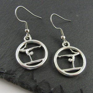 May include: Silver earrings featuring a gymnast silhouette on a balance beam within a circular design.