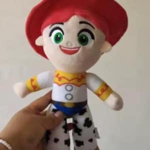 May include: A plush doll featuring a red cowboy hat, green eyes, and a smiling expression. The doll wears a white shirt with yellow accents and two circular buttons, paired with white pants featuring black cow-print spots and a blue belt.