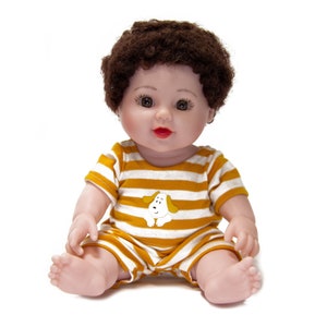 Puede incluir: Una muñeca bebé de pelo castaño y ojos marrones que lleva un mono a rayas amarillas y blancas con un gráfico de un cachorro blanco.