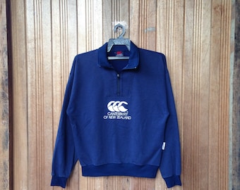 Puede incluir: Una sudadera azul marino con un logotipo blanco de Canterbury de Nueva Zelanda. La sudadera tiene una cremallera de media cremallera y un cuello de canalé.