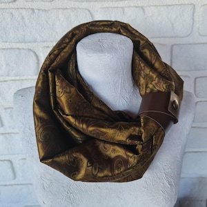 May include: A gold and brown paisley patterned infinity scarf with a brown leather cuff. The scarf is draped over a white mannequin head. The scarf is made of a soft, flowing fabric.